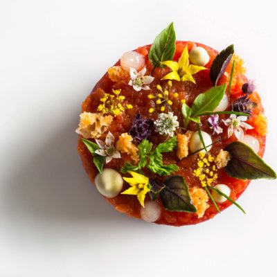 Tomato lobster disc garnished with croutons, chervil, chive tips, lemon gel, bonito mayonnaise, tomato flowers, fennel flowers, and basil on a white surface. Prepared by Daniel Humm at Eleven Madison Park.