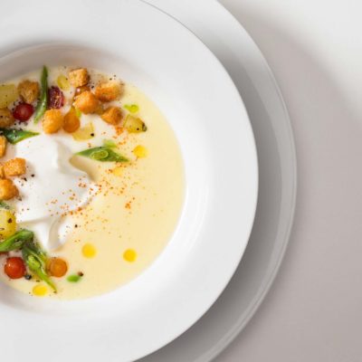 Cold potato and leak soup with yogurt, green beans, potatoes, currant tomatoes, croutons, extra virgin olive oil and hot pepper. Served on a white ceramic bowl on a white background.